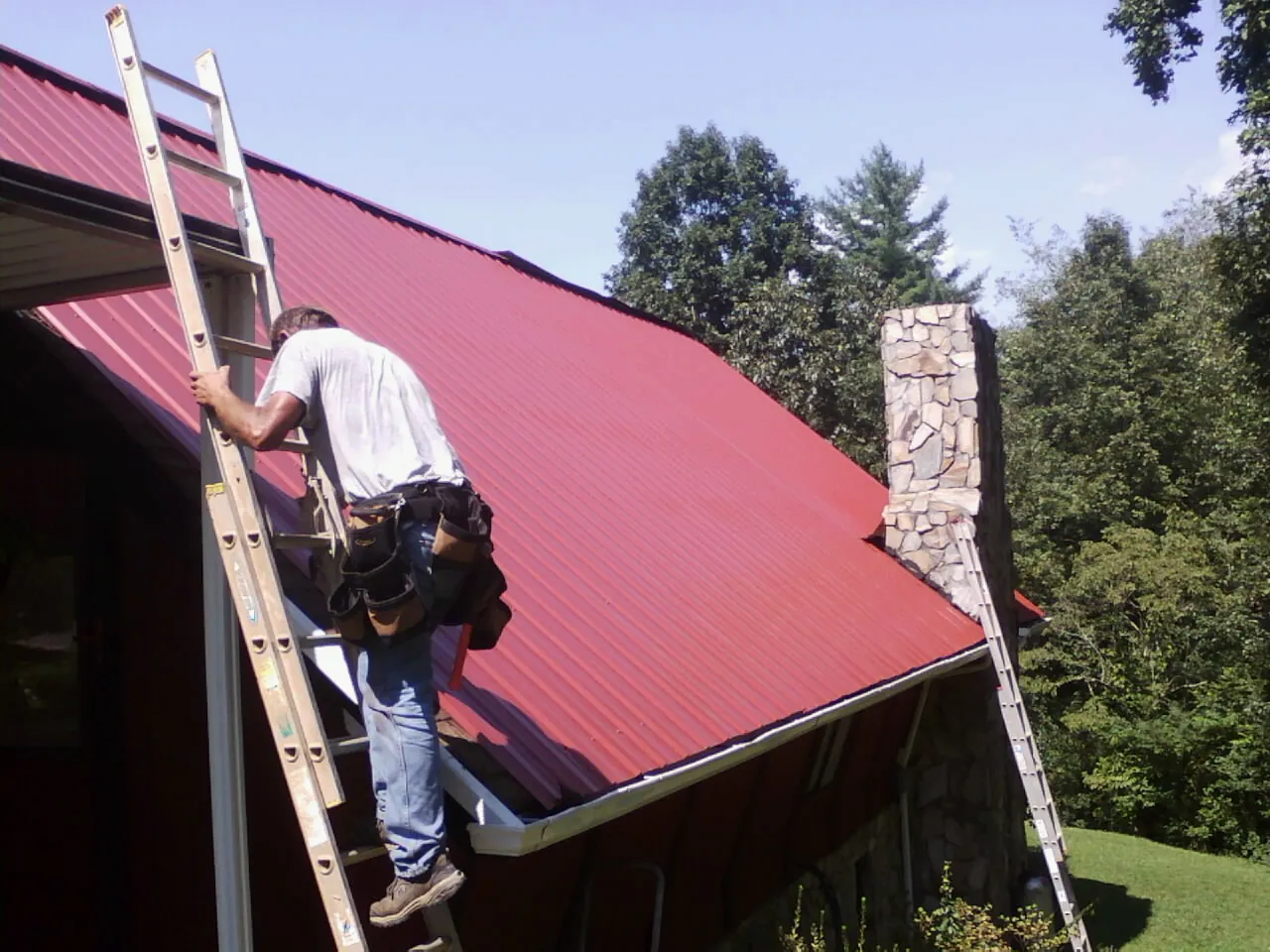 Roofer on a ladder beside a red metal roof and stone chimney — Soffit & Fascia Repair by Roofing Brothers in New Knoxville